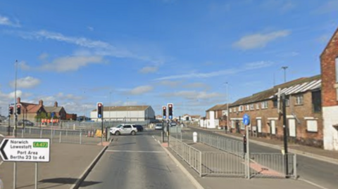 A pedestrian crossing. To the right, a pedestrian island separates the carriageway. To the left, a pedestrian island leads to another set of traffic lights further to the left. A white road sign pointing left states Norwich Lowestoft A47, Port Area Berths 23 to 44. Industrial buildings are in the background.