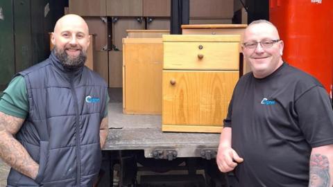 Two men, standing by an open lorry with lots of items inside, including cabinets and a red wheelie bin. The man on the left is wearing a blue gilet, with a green T-shirt, and the man on the right has on a black T-shirt. They are both smiling and looking straight at the camera.