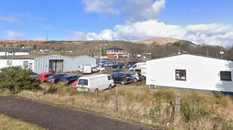 A small industrial estate with cars and vans parked in the car park. It is surrounded by grass with hills far in the background