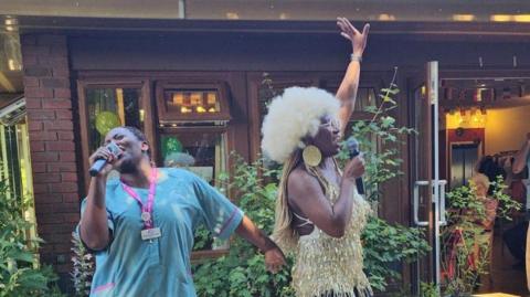 Staff at Abbey Park care home entertain residents outside the main building in the shade. A nurse on the left of the picture is wearing a light blue uniform smock and singing into a microphone. Her colleague is wearing a gold dress with a blonde wig, similar to the look of Motown legend Tina Turner.