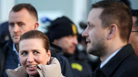 Danish PM Mette Frederiksen, wearing a puffer jacket with the collar up, smiles and looks towards Greenland PM Jens-Frederik Nielsen, who stands in profile at the right of the picture.