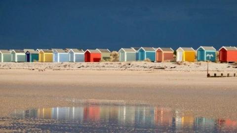 Beach huts on a beach in Moray. The sky above is dark blue. The sand in the foreground is gold and there is water in the extreme foreground. The huts are several different colours.