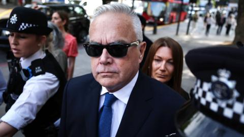 Former 鶹ҳ news presenter Huw Edwards, wearing a navy suit, blue tie and sunglasses, is flanked by police officers outside a London court in July