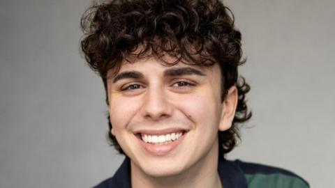 Sonny Monaghan. Sonny's a young man with dark curly hair and brown eyes. He smiles at the camera, wearing a blue and green striped polo shirt. 