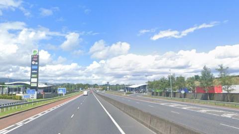 The Belfast-bound lanes of the M1 - there is a service station to the left. The sky is blue with white clouds.