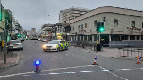 A photo of the scene on Southchurch Road in Southend-on-Sea in Essex. The photo shows the road cordoned off with police cones with blue lights on them. A police car can be seen within the cordon and there is police tape blocking off the pavement. 