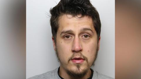 A man with dark hair and a goatee beard wearing a police issue grey sweatshirt 