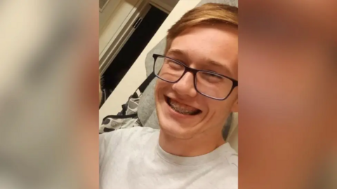 Ryan Marshall, a teenage boy, smiling widely at the camera. He has blonde short hair in a side parting, wears black framed square glasses and has braces and is wearing a light grey round neck t shirt.