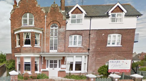 Friary House is a detached, three-storey, brick building behind a low wooden fence.