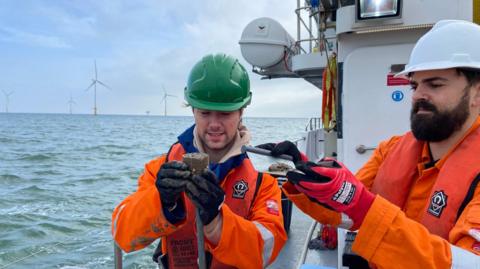 Two researchers dressed in orange waterproofs, the man on the left wearing a green hard had and the man on the right wearing a white hard hat, each lifting up and looking at seabed samples, on a white boat with the sea and windfarm turbines behind them 