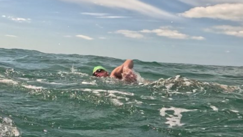 Man swimming in the Channel. He is wearing a green swimming hat and black goggles and the water is a green/blue. The sky is blue with white clouds. There's a blue boat which he is swimming alongside.