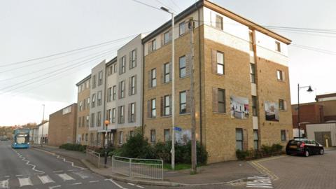A four-storey accommodation block on a street in day time. There is a bus in the distance approaching a zebra crossing and a parked car to the right of the building.