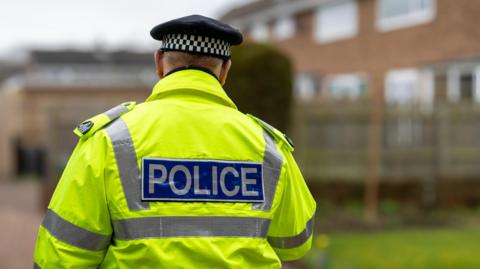 A photo of a police man with his back to the camera in a high vis coat with 'police' written on the back. He has a black police hat on with black and white checked markings on. The background is blurred but shows him walking towards some residential flats. 