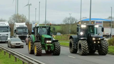 Two tractors drive down a dual carriageway as traffic starts to build up behind them. 