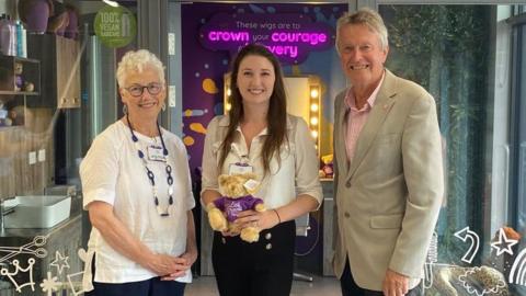 Dr Catherine Pointer holding a small teddy bear with LPT Trustee Tim Lowe and Dame Kathy August, outside a shop front with purple and white displays in the window
