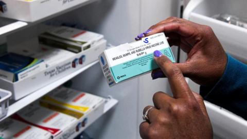 A box containing the vaccine against measles, mumps and rubella, being held by a woman with her right hand, with the index finger of the left pointing to the writing on it.