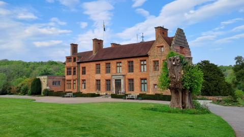 The image shows a large country house, with red brick and a flag raised on its roof. Trees can be seen in the background as well as a bright green lawn. A few clouds can be seen in the sky.