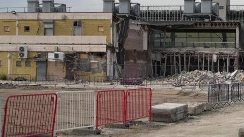 Image shows Stretford Mall in the process of being demolished. It is a late 1960s building with much of one facade now removed and the interior exposed. 