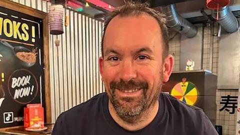 A man in his early 40s, with short dark hair and a short beard. He appears to be sitting in a restaurant and is smiling into a camera.
