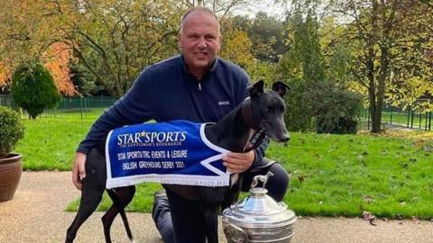 A dog trainer behind a greyhound wearing a winner sash in a field.