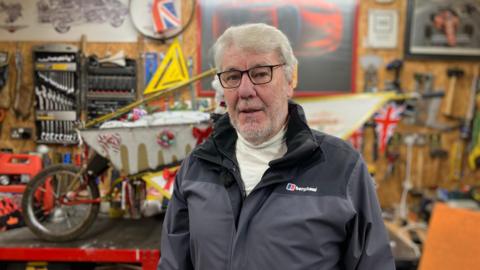 A man in his 70s stands in front of a wheelbarrow, adorned in Christmas decorations including bows and baubles, in his garage. There are tools neatly hung all over the wall. He is wearing a white turtleneck, a black and grey coat, and black-rimmed glasses.