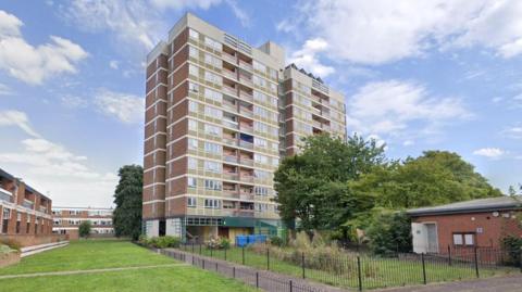 A screenshot from Google street view showing the existing residential tower block. It is a tall brick building with balconies on each floor and a fenced green space at the bottom.