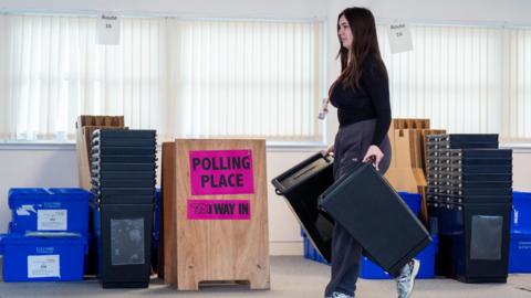 Woman walks with black election ballot boxes past a sign saying "polling place, this way" and a number of crates