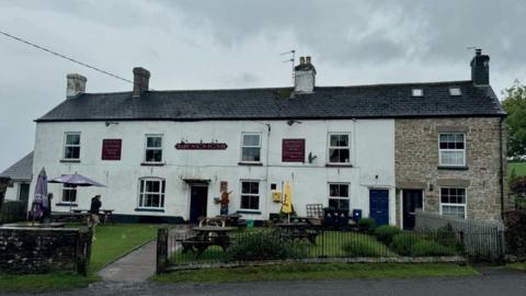 Two storey pub, with four well-spaced windows on each floor. There is a grass garden in front with round benches and a black metal fence. A small red and gold sign above the door reads The Nag's Head