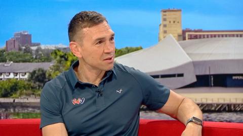 A man with short brown hair wearing a blue polo shirt sits on a red sofa in a TV studio.