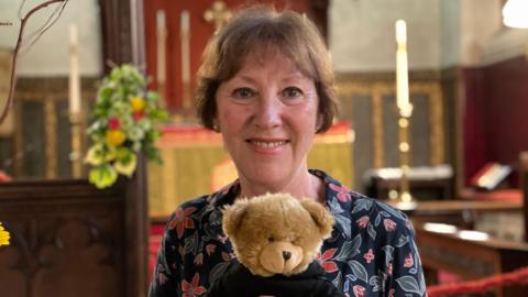 Helen Jones wearing a blue floral dress holding a teddy bear. She is inside a church and the altar is visible behind her draped with a gold and red cloth.