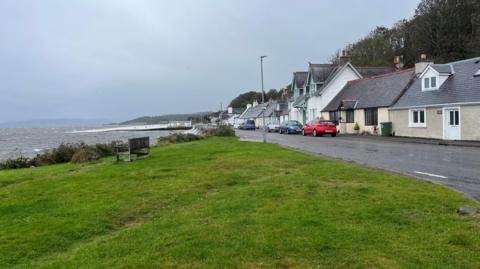 Houses line the street along the waterfront at North Kessock. It's a wet day and the Moray Firth looks grey and choppy. There is a grassy bank in the foreground, and the old ferry pier is visible in the background.