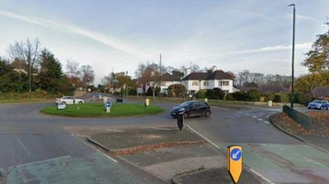 A roundabout with two cars driving around it - one white and one black. Houses can be seen on the other sides of the road.