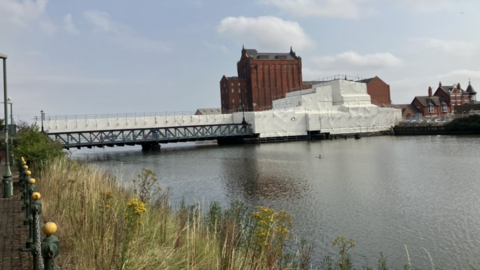 Corporation Bridge covered in white sheeting spanning a calm river. There is a very tall brick building in the background and the sky is blue with a few clouds. There are reeds in the foreground