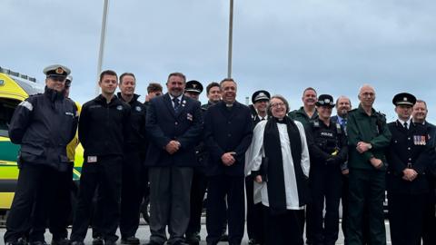 Men and women in different uniforms, including police, fire, paramedics and ecclesiastical, stand with hands clasped in front of two flag poles.