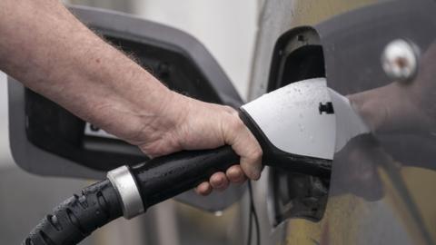 A hand holding a white electric vehicle charger plugged into a car. It is raining in the image.