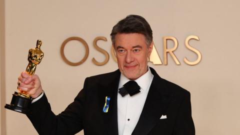 Peter Straughan poses with the Best Adapted Screenplay for "Conclave" award in the Oscars photo room at the 97th Academy Awards. He is wearing a black suit and has blue and yellow ribbons pinned to his lapel.