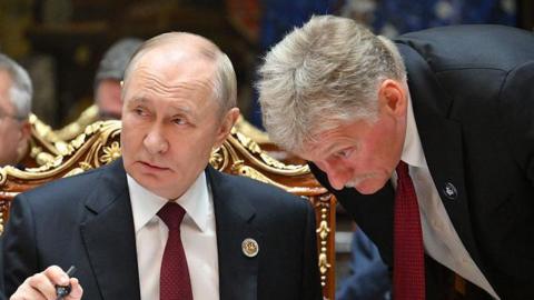Russian President Vladimir Putin in a suit and red tie say behind a table, Peskov, a man with grey hair wearing a suit is bending down listening to him talk.