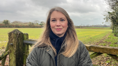 Laura Layzell wearing a green coast with a navy blue top and navy blue scarf. She has long dark blonde hair and is smiling at the camera. Behind her is a field with a wooden gate just before it.