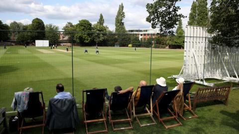 Guests sitting in deckchairs watch a cricket match from behind a black net. A white sight screen is to the right and players can be seen gathered around the batter on a brown cricket strip surrounded by green grass