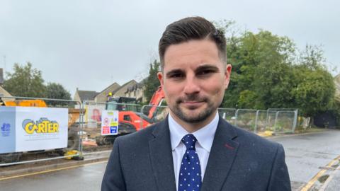 Joe Harris, a man with neat brown hair and a brown beard, stands on the pavement beside a wet road, with a construction site and houses behind him.