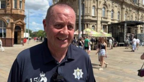 A man is smiling and wears a dark blue top with regimental logos. Various stalls and buildings can be seen in the background.