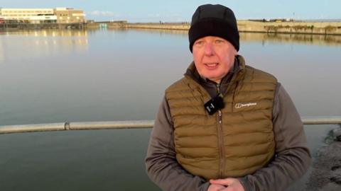 Colin Clayton, who chairs the group, is standing in front of the marine lake. He is wearing a black hat, grey jumper and khaki gilet.