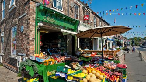 A lively street market scene outside a green-painted shop called “Millie’s,” with tables and crates full of colourful fruits and vegetables such as melons, apples, pineapples, and tomatoes. A beige umbrella shades the produce, while festive bunting hangs across the street. Shoppers stroll past other nearby cafés and shops under a bright blue sky.