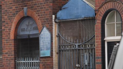A close-up of a red brick building featuring an arched entrance with a Star of David and the years 1923 and 5684 inscribed above the door. A blue sign for the "East London Central Synagogue" is mounted on the wall. To the right of the entrance is a tall, padlocked metal gate topped with a weathered blue gabled roof.