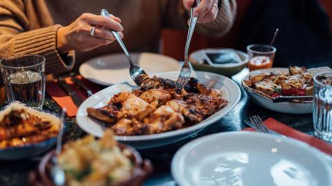 A person is using two forks to lift meat out of a dish in the middle of a restaurant table, surrounded by other smaller dishes