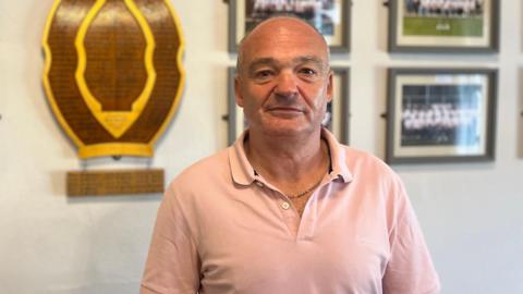 Neville Warnes is looking directly at the camera and smiling. He is wearing a pale pink short-sleeved polo shirt and a thin chain round his neck.