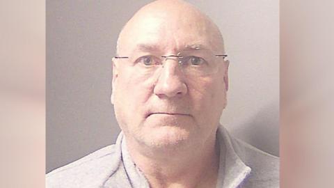 Custody image of Michael - wearing a grey collar jumper and glasses- looking into the camera 