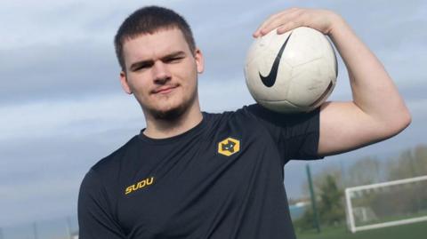 A male teenager in a black shirt with a yellow Wolves FC badge holding a football on his shoulder. He is standing on an astroturf pitch with a goal in the background.