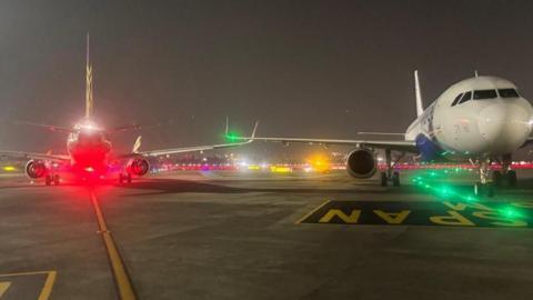 Two airplanes facing in the opposite directions in adjacent taxiways in an airport at nighttime, with the tip of their right wings in contact