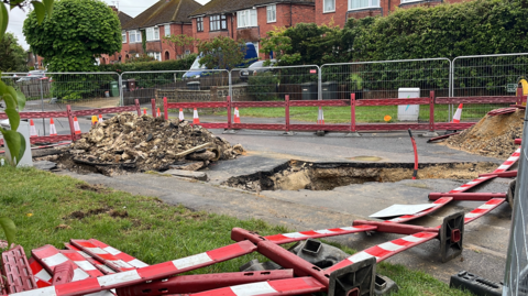 A road with a five foot trench and rubble on the road which is all fenced off and a red wire sticking out of the trench
